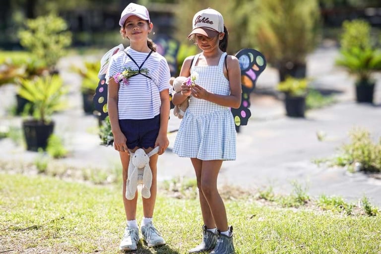Two children wearing white caps and roller skates stand together on a sunny outdoor path surrounded by plants