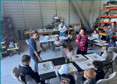 Group of children and adults in an art workshop inside a warehouse studio with tables, supplies, and storage shelving