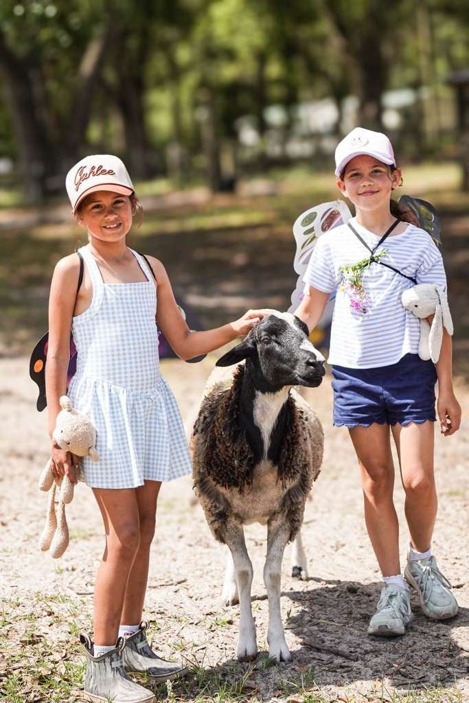 Two young girls in white caps and summer clothing walk with a gray and white goat on a tree-lined path