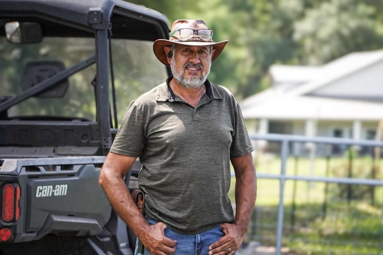 Middle-aged man in a cowboy hat and work clothes standing beside a black Can-Am utility vehicle on a ranch with buildings visible in the background