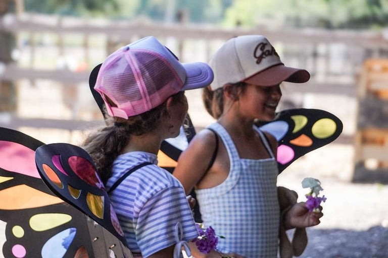 Two children wearing colorful butterfly wings and caps, standing outdoors in an urban setting