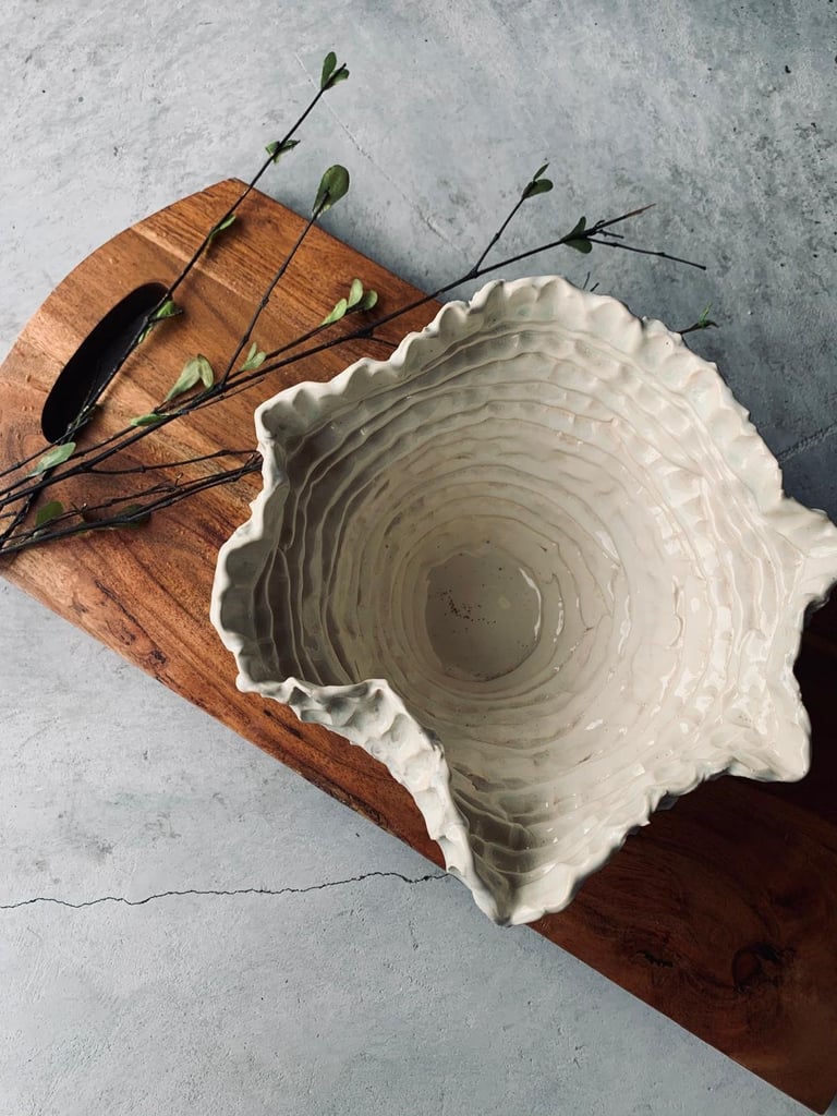 White ceramic shell-shaped dish on wooden board with dried botanical stems and flowers on gray surface