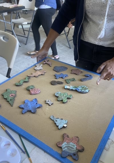 Group displaying colorful handmade ceramic and clay ornaments on a tan board with blue border at a community gathering