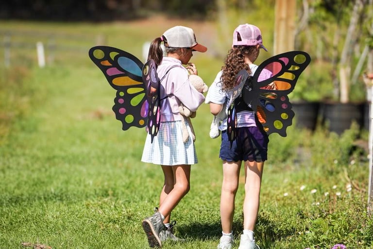 Two children wearing colorful butterfly wings and caps run through a grassy outdoor field on a sunny day