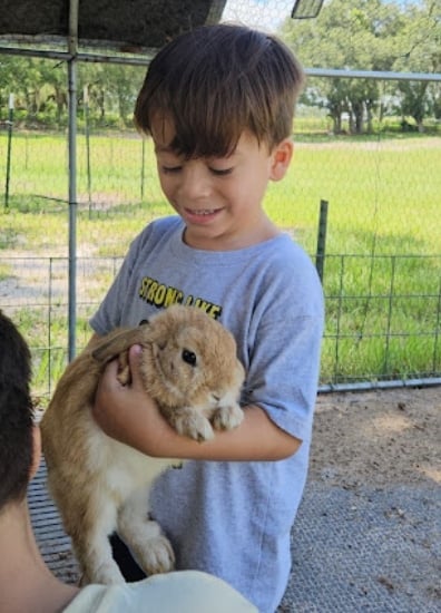 Young boy smiling while holding a brown rabbit in his arms outdoors with a farm fence in the background