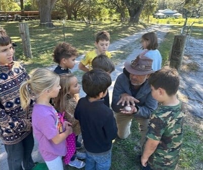 A group of children gathered around an adult in a cowboy hat on a rural property with wooden fencing and trees