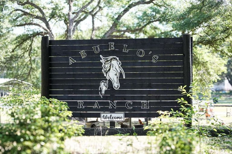 Black wooden sign reading Abrillos Ranch with a horse illustration and welcome message, surrounded by trees and greenery