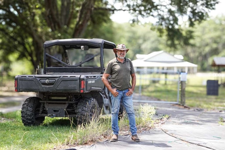Man in cowboy hat standing next to black utility vehicle on tree-lined driveway with house in background