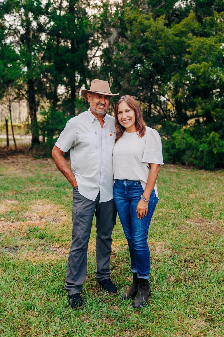 A man and woman standing together on grass with trees in the background. The man wears a cowboy hat and light blue shirt, while the woman wears a cream t-shirt and blue jeans.